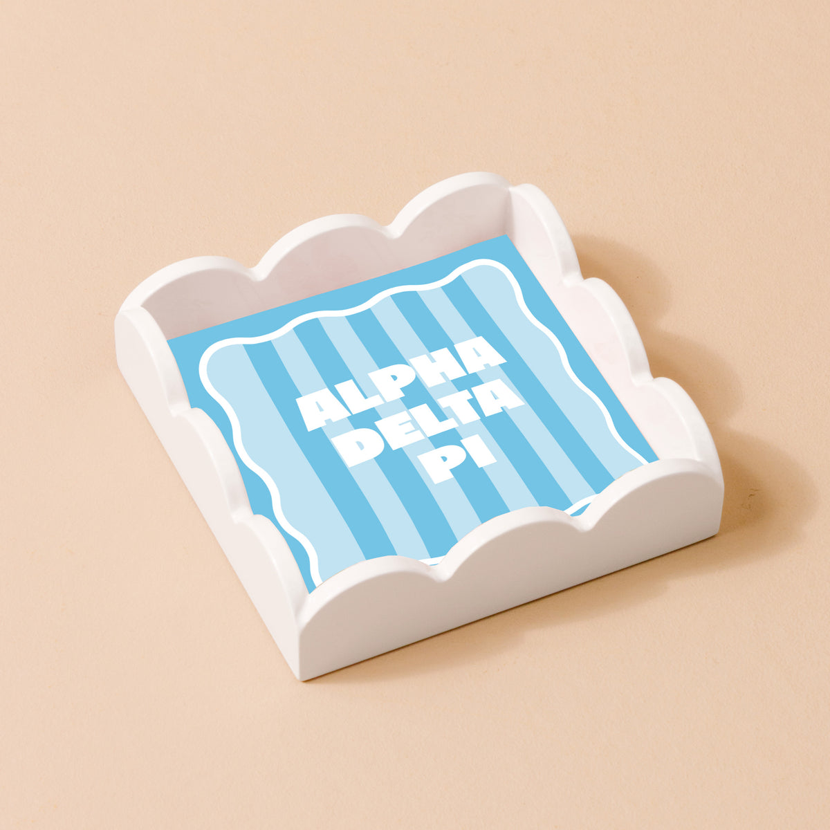 The Darling Effect’s Alpha Delta Pi Scallop Trinket Tray, shown holding a blue striped notepad with bold white "ALPHA DELTA PI" text, is a stylish beige desk accessory perfect for organizing essentials.
