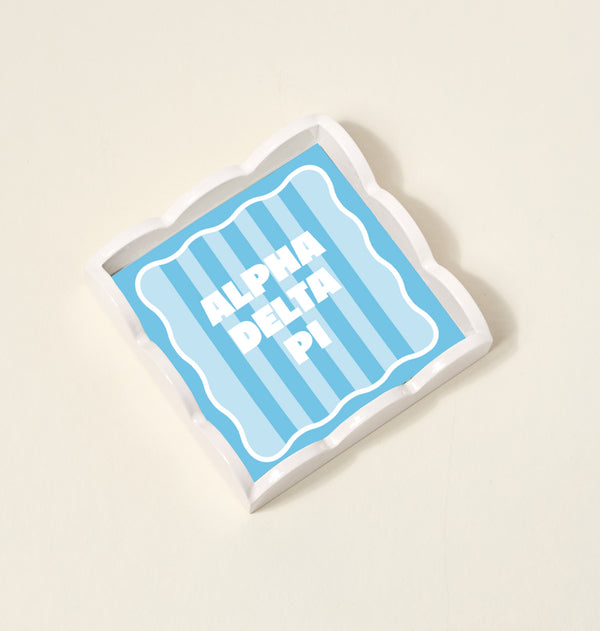 The Darling Effect Alpha Delta Pi Scallop Trinket Tray features a square shape with blue and light blue vertical stripes and bold white ALPHA DELTA PI text, perfect as a stylish jewelry organizer or accessory tray.