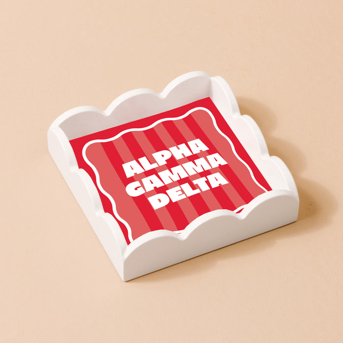 The Darling Effect's Alpha Gamma Delta Scallop Trinket Tray features a scalloped white design holding a red and pink striped square with bold ALPHA GAMMA DELTA lettering—ideal for jewelry or keepsakes.