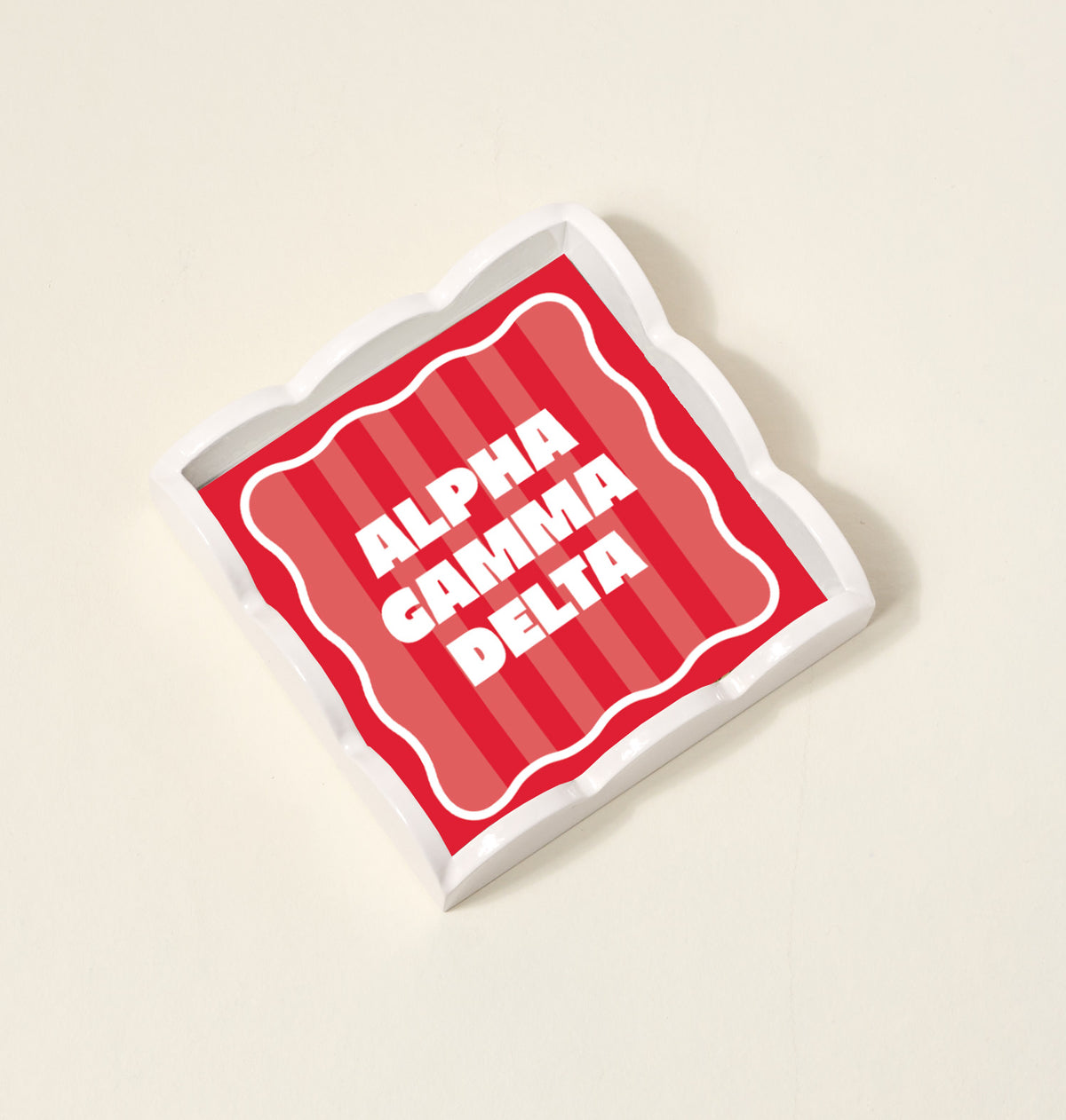 The Darling Effect’s Alpha Gamma Delta Scallop Trinket Tray is a square ceramic tray with wavy edges, red and pink stripes, and ALPHA GAMMA DELTA in white at the center—perfect for storing jewelry or accessories.