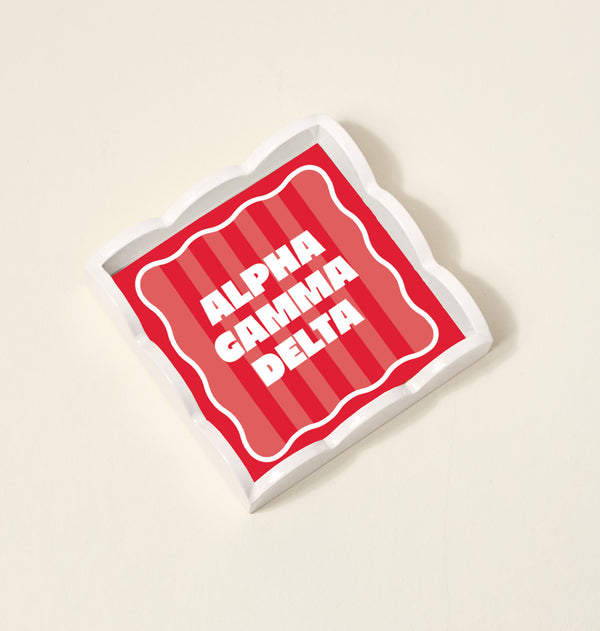 The Darling Effect’s Alpha Gamma Delta Scallop Trinket Tray is a square ceramic tray with wavy edges, red and pink stripes, and ALPHA GAMMA DELTA in white at the center—perfect for storing jewelry or accessories.