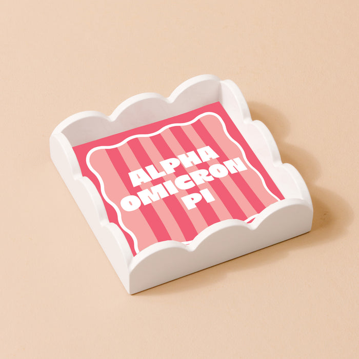 The Darling Effect Alpha Omicron Pi Scallop Trinket Tray features a white finish with a pink and red striped base and bold white lettering—ideal as an accessory tray or jewelry organizer.