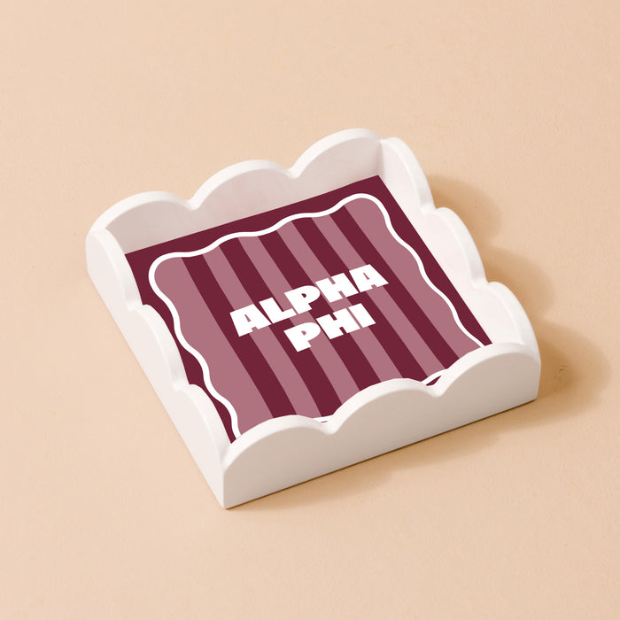The Darling Effect’s Alpha Phi Scallop Trinket Tray features a white scallop design with a square insert of maroon and pink stripes and bold white ALPHA PHI text—a stylish jewelry organizer or accent tray for any space.