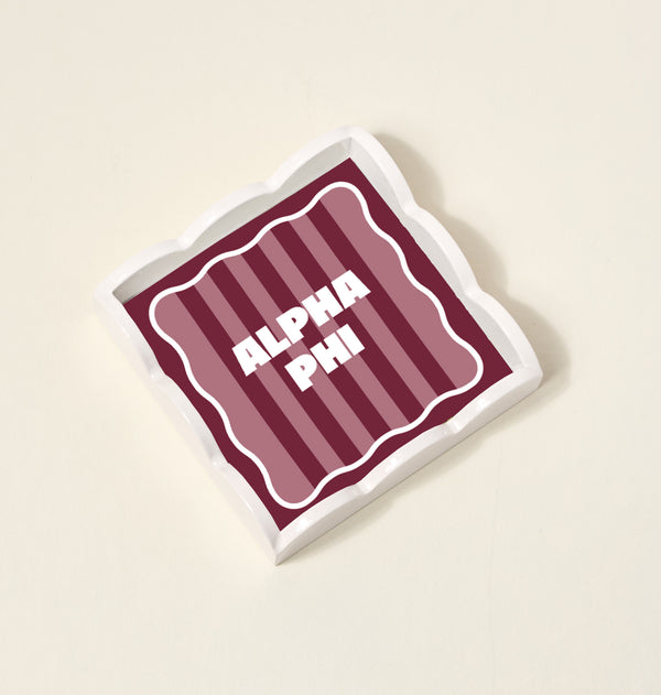 The Darling Effect’s Alpha Phi Scallop Trinket Tray features a square ceramic design with wavy edges, maroon and pink stripes, and bold white ALPHA PHI lettering—ideal as a chic jewelry organizer or accessory tray.