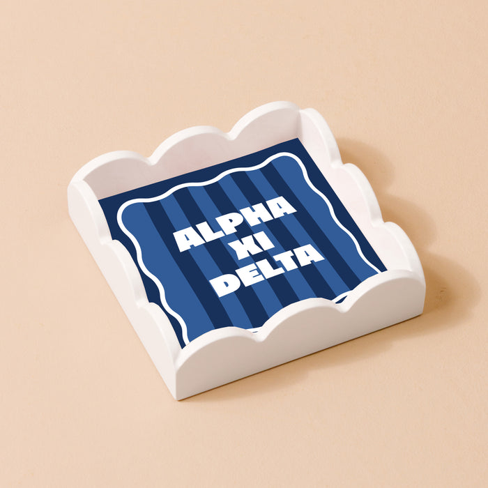 The Darling Effect’s Alpha Xi Delta Scallop Trinket Tray pairs stylishly with a blue ALPHA XI DELTA napkin and striped backdrop, making it an ideal chic jewelry organizer or vanity tray.