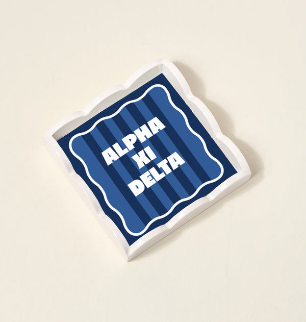 The Darling Effect's Alpha Xi Delta Scallop Trinket Tray features a blue striped design with bold white ALPHA XI DELTA lettering—ideal as a stylish vanity tray or jewelry organizer.