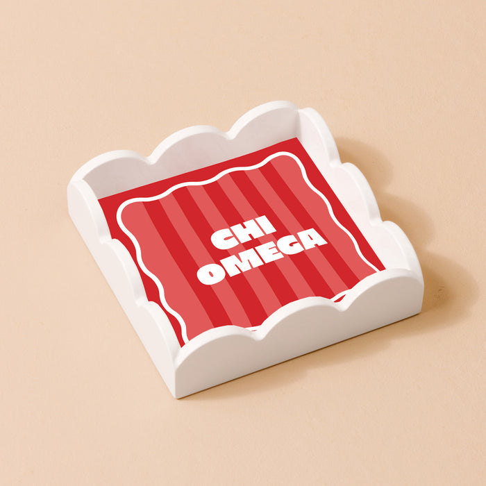 The Darling Effect’s Chi Omega Scallop Trinket Tray features a white scallop design with a red striped inset and bold white CHI OMEGA text, perfect for organizing jewelry or use as a decorative accent.