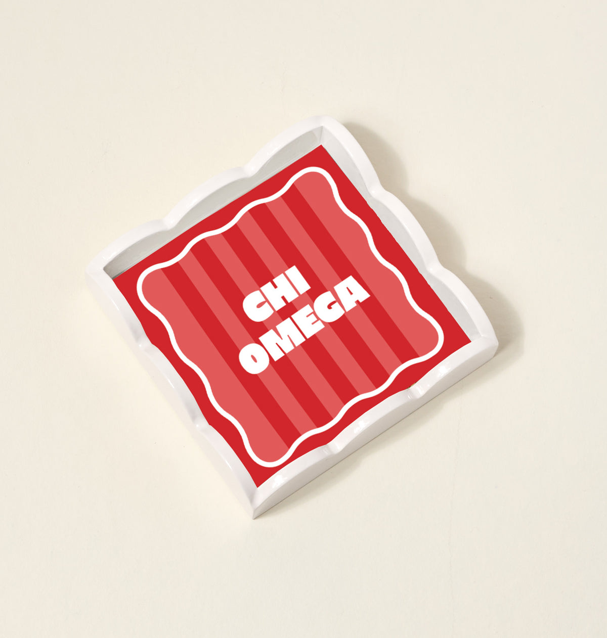 The Darling Effect’s Chi Omega Scallop Trinket Tray is a square ceramic tray with wavy edges, a red striped background, and bold white CHI OMEGA lettering—perfect as a jewelry organizer or decorative accent.