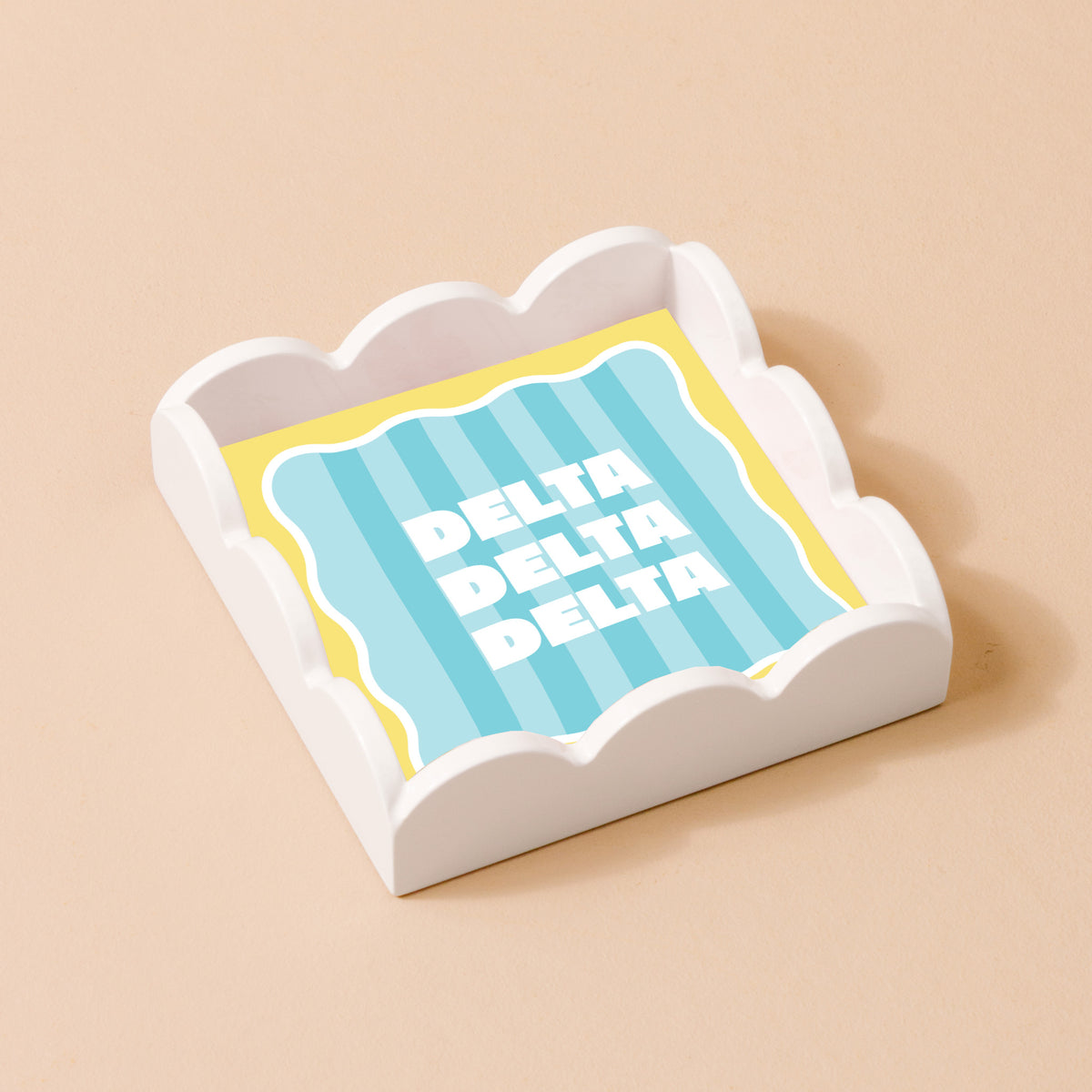 The Darling Effect’s Delta Delta Delta Scallop Trinket Tray holds a square napkin with blue and yellow stripes and “DELTA” printed three times in bold white letters, set on a light beige background.