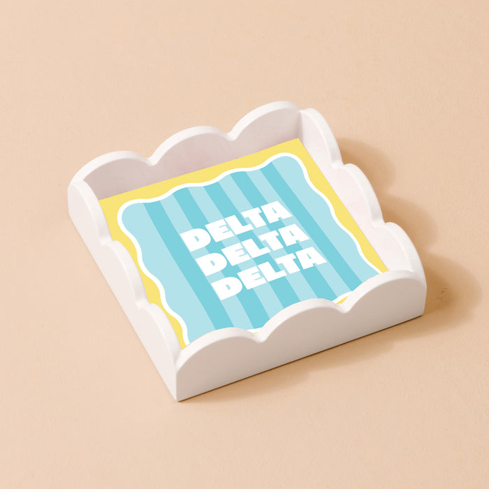 The Darling Effect’s Delta Delta Delta Scallop Trinket Tray holds a square napkin with blue and yellow stripes and “DELTA” printed three times in bold white letters, set on a light beige background.