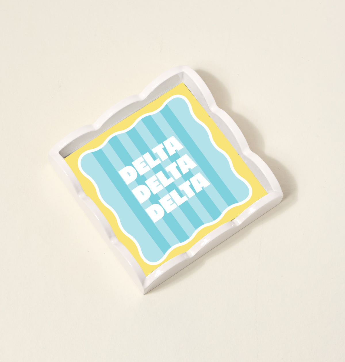 The Darling Effect Delta Delta Delta Scallop Trinket Tray features a square, scalloped-edge design with blue and yellow stripes and bold white lettering—perfect as a jewelry organizer or accessory tray.