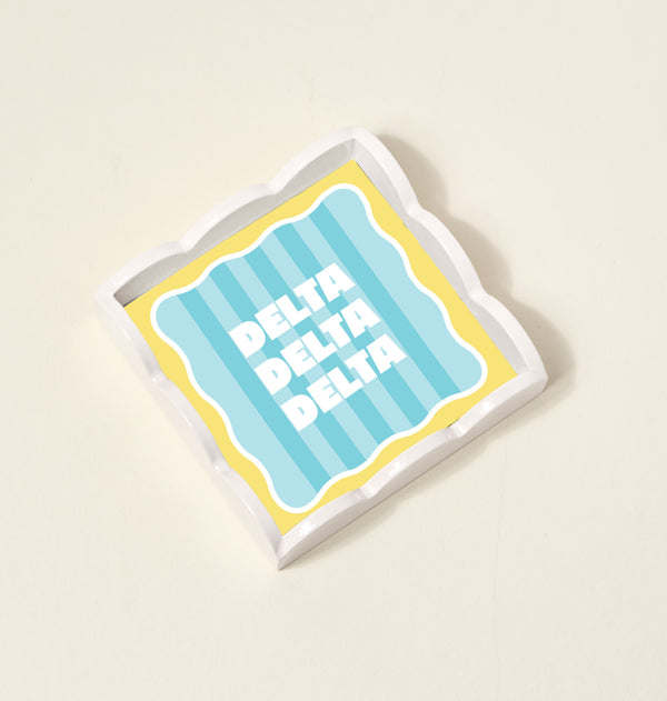 The Darling Effect Delta Delta Delta Scallop Trinket Tray features a square, scalloped-edge design with blue and yellow stripes and bold white lettering—perfect as a jewelry organizer or accessory tray.