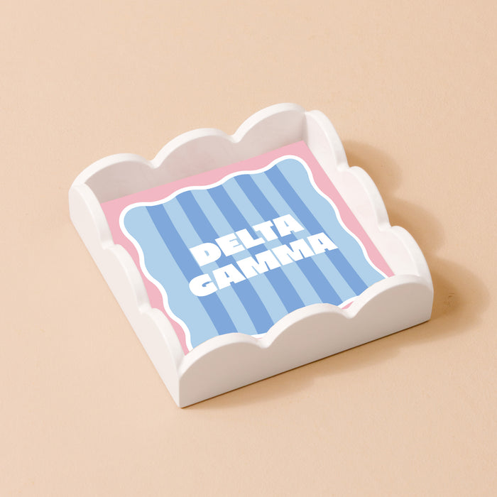 The Darling Effect’s Delta Gamma Scallop Trinket Tray features a white scalloped design holding a square card with blue stripes, a pink border, and bold DELTA GAMMA lettering. This stylish accessory tray sits on a beige surface.