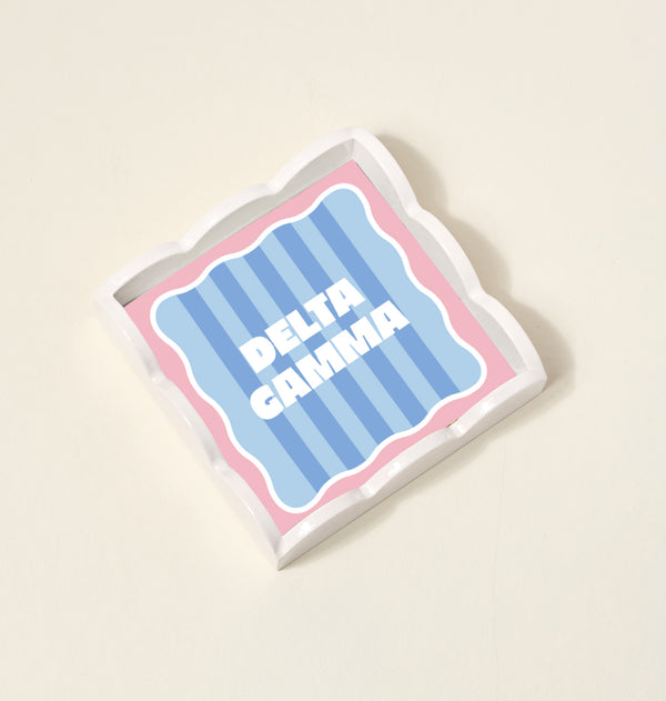 The Darling Effect Delta Gamma Scallop Trinket Tray features blue stripes, a pink border, and wavy edges with bold white DELTA GAMMA lettering—ideal as a jewelry organizer or accessory tray.
