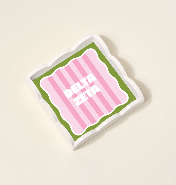 The Darling Effect’s Delta Zeta Scallop Trinket Tray features a square shape, green wavy border, pink striped center, and bold white DELTA ZETA letters—ideal as a jewelry organizer or vanity tray. Shown on a plain beige background.