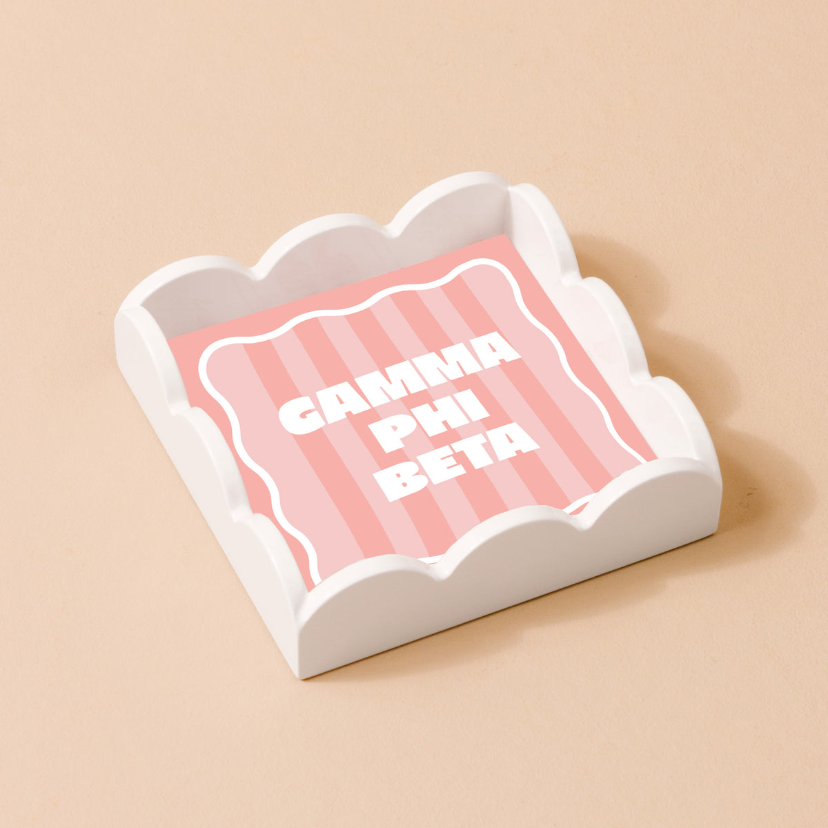 The Darling Effect’s Gamma Phi Beta Scallop Trinket Tray features pink and white stripes with bold white GAMMA PHI BETA letters—perfect as a jewelry holder or vanity accent.