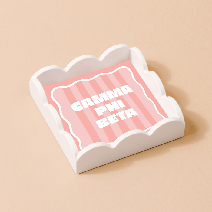 The Darling Effect’s Gamma Phi Beta Scallop Trinket Tray features pink and white stripes with bold white GAMMA PHI BETA letters—perfect as a jewelry holder or vanity accent.