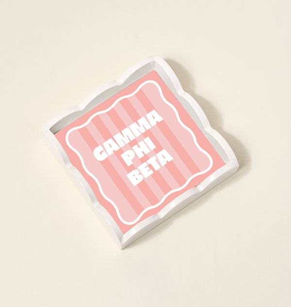 The Darling Effect Gamma Phi Beta Scallop Trinket Tray, featuring a wavy edge, pink striped background, and bold white "GAMMA PHI BETA" lettering at the center, is displayed on a light surface.