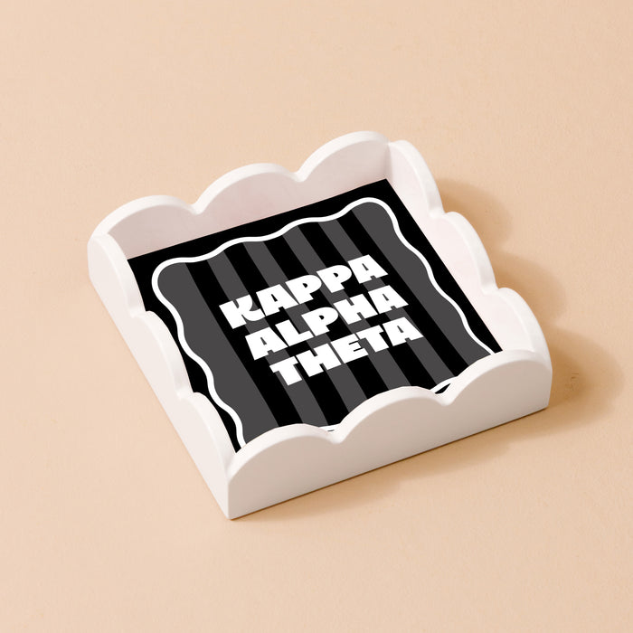 The Kappa Alpha Theta Scallop Trinket Tray by The Darling Effect rests on a beige surface, featuring a black and gray striped insert with bold white text—ideal as a chic organizer for jewelry or accessories.