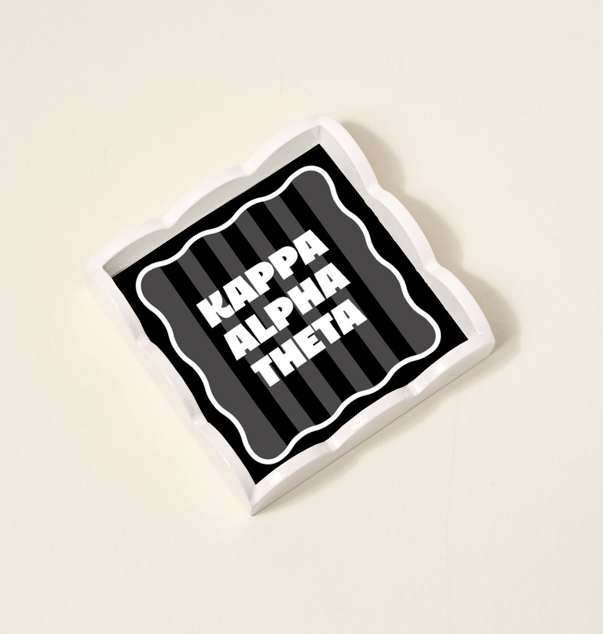 The Kappa Alpha Theta Scallop Trinket Tray by The Darling Effect features bold white text on a black background with dark gray stripes and a wavy edge—ideal as an accessory tray or jewelry organizer.