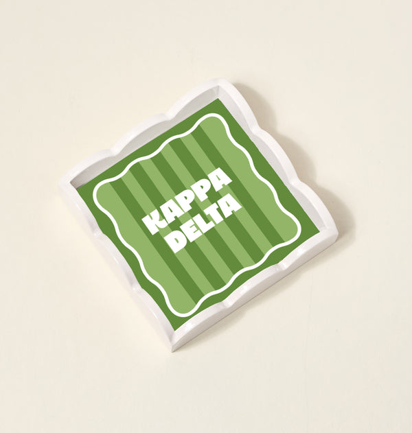 The Darling Effect's Kappa Delta Scallop Trinket Tray features a green striped, wavy-edged square design with bold white KAPPA DELTA lettering—ideal as an accessory or jewelry organizer.
