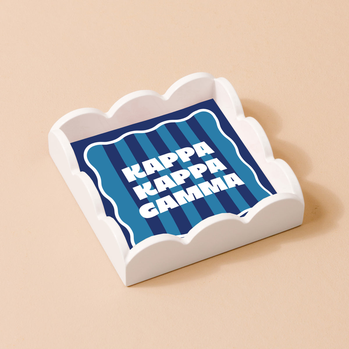 The Darling Effect’s Kappa Kappa Gamma Scallop Trinket Tray features “KAPPA KAPPA GAMMA” in bold white letters on a blue striped background, displayed on a light beige surface.