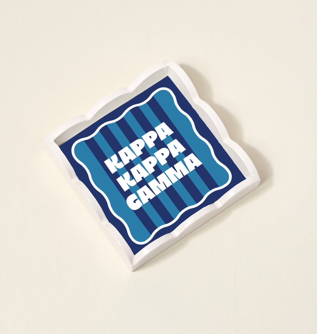The Darling Effect Kappa Kappa Gamma Scallop Trinket Tray features wavy edges, blue and navy stripes, and bold white lettering—perfect as a vanity tray or jewelry organizer.