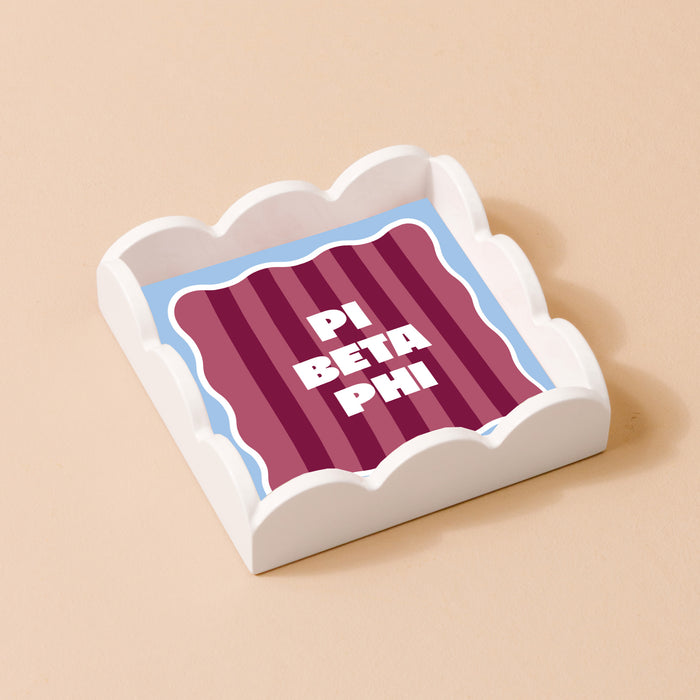 The Darling Effect’s Pi Beta Phi Scallop Trinket Tray features maroon and pink stripes with “PI BETA PHI” in bold white letters, perfect as a jewelry organizer or decorative accent.