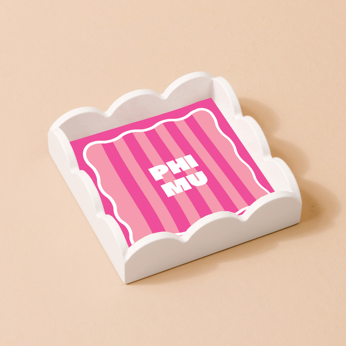 The Phi Mu Scallop Trinket Tray by The Darling Effect features a white scalloped design holding a pink card with bold white PHI MU lettering, perfect as an accessory or jewelry organizer. Displayed on a light beige surface.