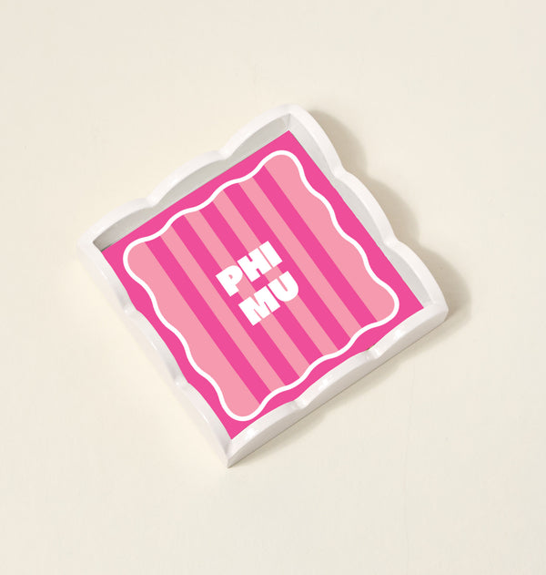 The Darling Effect's Phi Mu Scallop Trinket Tray features pink and light pink vertical stripes, bold white PHI MU lettering at the center, a scalloped edge, white border, and sits on a light beige surface.