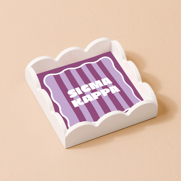 The Darling Effect Sigma Kappa Scallop Trinket Tray is white with a purple striped base and bold white "SIGMA KAPPA" letters—perfect as a stylish accessory tray.