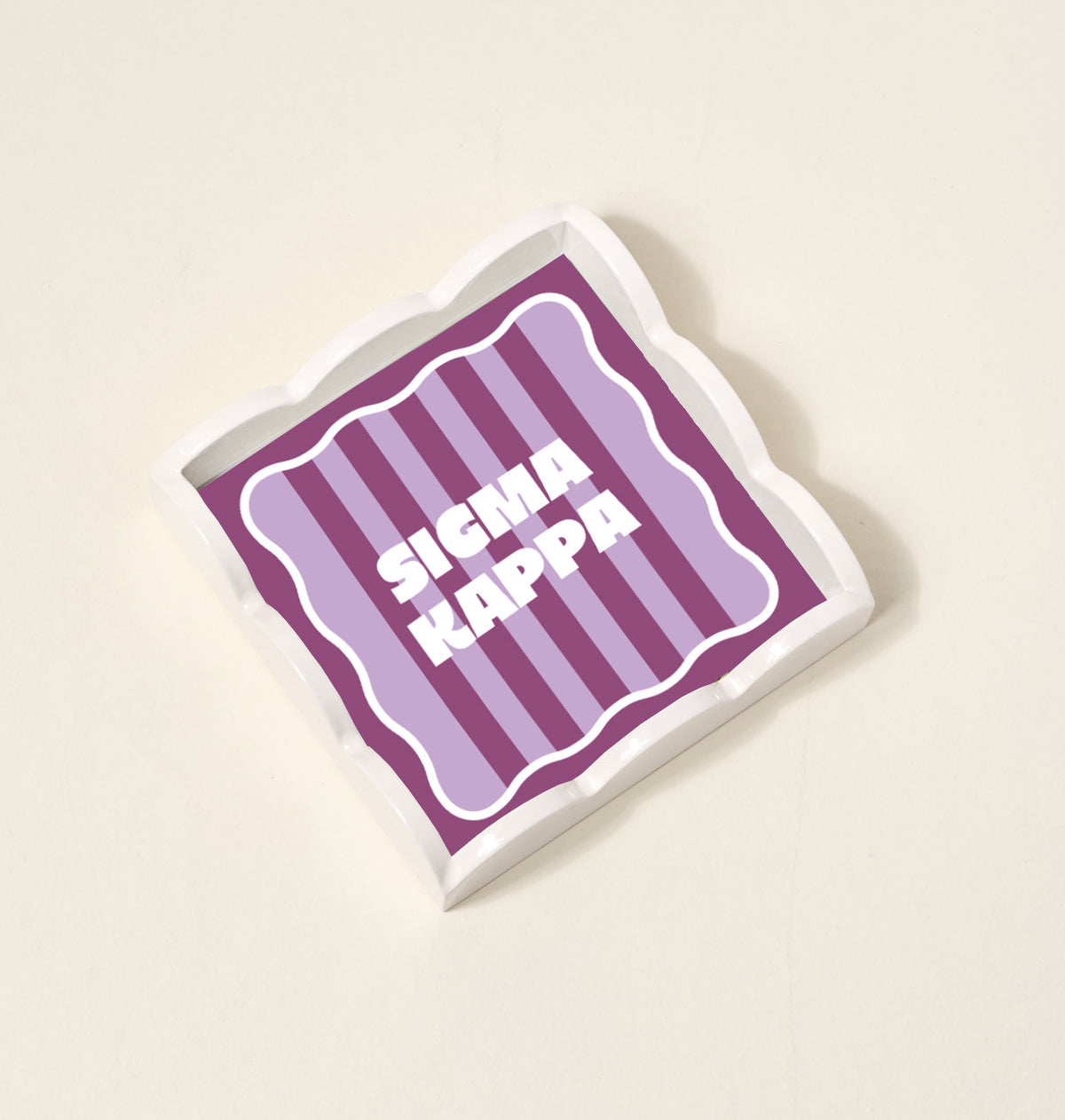 The Darling Effect’s Sigma Kappa Scallop Trinket Tray features wavy edges, purple and pink vertical stripes, and bold white “SIGMA KAPPA” text—perfect as an accessory tray or jewelry organizer.