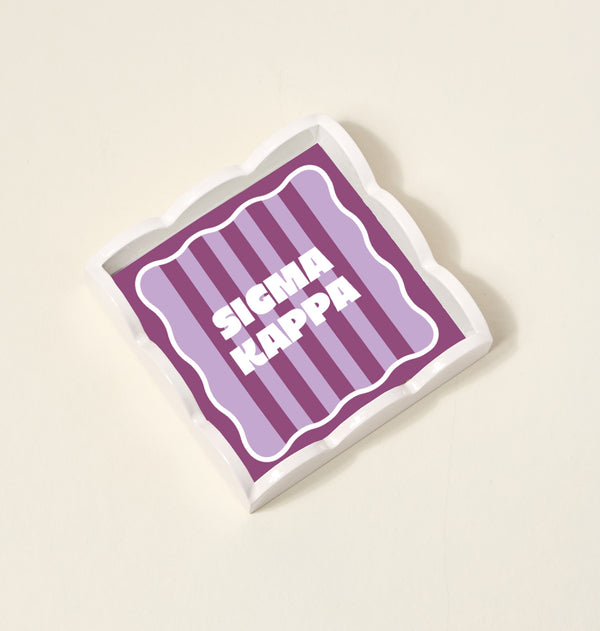 The Darling Effect’s Sigma Kappa Scallop Trinket Tray features wavy edges, purple and pink vertical stripes, and bold white “SIGMA KAPPA” text—perfect as an accessory tray or jewelry organizer.