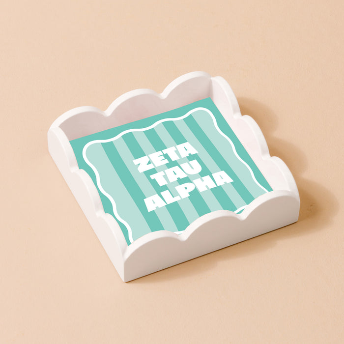 The Darling Effect's Zeta Tau Alpha Scallop Trinket Tray features a white scallop design with a mint green and white striped base and bold white ZETA TAU ALPHA letters, ideal as an accessory or jewelry organizer against a beige background.