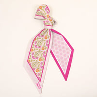 The Darling Effect's Chi Omega Twirl & Tie Scarf Accessory is a silk scarf with a pink, yellow, and green floral pattern on cream, pink border, and OX in pink. Perfect for versatile styling and sorority spirit.