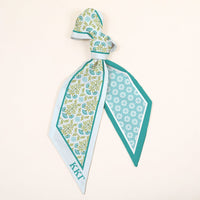 The Darling Effect’s Kappa Kappa Gamma Twirl & Tie Scarf Accessory features blue and green floral and geometric patterns, light blue edges, a KKG monogram, and instantly adds boutique charm to your look.