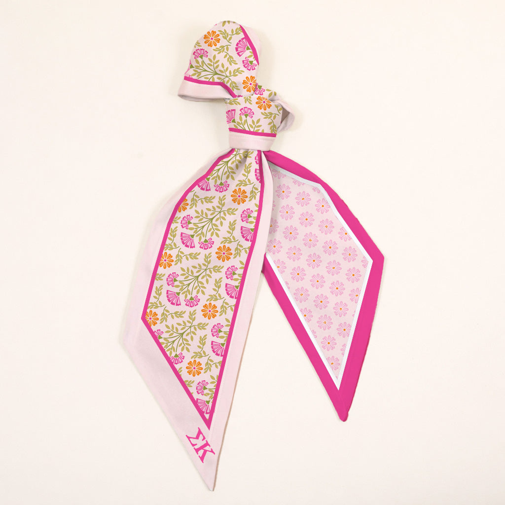 The Darling Effect's Sigma Kappa Twirl & Tie Scarf Accessory features pink, yellow, and green floral print on one side, a pink geometric pattern on the other, and bright pink edging—perfect for versatile styling on a cream background.
