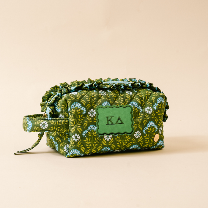The Darling Effect’s Kappa Delta Essentials Quilted Cosmetic Pouch features a green floral pattern, zippered closure, handle, ruffled trim, and a front patch with the Greek letters KΔ—perfect for carrying your sorority essentials in style.