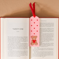 An open book shows chapter twenty-one with The Darling Effect’s Teddy Bear Hugs Acrylic Bookmark—decorated with a teddy bear, hearts, and a velvet ribbon tassel—resting on the pages, making an adorable romantic gift on a beige surface.