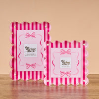 Two Sweetheart Stripes Rectangle Scalloped Acrylic Frames by The Darling Effect, featuring pink and red stripes with bow designs, are displayed on a wooden surface against a plain beige background.