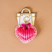 The Darling Effect's Heartthrob Puffy Heart Bag Charm features pink stripes, ruffled edges, a small pink bow, and gold hardware—an adorable Valentine’s Day accessory shown on a beige background.