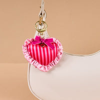 A close-up of a white handbag featuring The Darling Effect’s Heartthrob Puffy Heart Bag Charm—pink, striped, heart-shaped with ruffles and a bow—hanging from the handle on a gold clasp, standing out against a plain beige background.