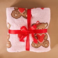The Darling Effect's Teddy Bear Hugs Cozy Dreams Luxe Blanket, featuring brown teddy bear and red heart motifs, is folded and tied with a red satin ribbon, evoking cozy warmth against a beige background.