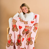 A smiling woman sits on a chair wrapped in The Darling Effect’s Teddy Bear Hugs Cozy Dreams Luxe Blanket, a pink ultra-soft microfiber throw with teddy bears holding red hearts and heart shapes, against a beige background.