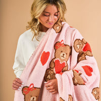 A blonde woman in a white top holds The Darling Effect’s Teddy Bear Hugs Cozy Dreams Luxe Blanket, an ultra-soft pink microfiber throw with teddy bears, red hearts, and bows, against a beige background.