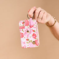 A hand holds The Darling Effect’s Rose Garden Keychain Wallet, featuring pink and red florals, gold hardware, a keyring, snap closure, and zipper pocket. The person wears a gold ring and beaded bracelet against a beige background.
