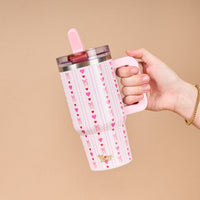A hand holds The Darling Effect’s Oh So Lovely 20 oz Lifestyle Flip Straw Tumbler, featuring pink hearts and bows on insulated stainless steel, shown against a white background.