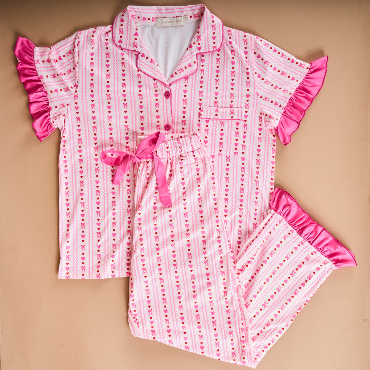 Perfect for a cozy night in, the Oh So Lovely Slumber Party Pajama Pants Set by The Darling Effect features a pink and white striped pajama set adorned with petite red hearts, pink buttons, a chest pocket, and whimsical pink ruffle trim on the sleeves and pant cuffs. Crafted from ultra-soft fabric for maximum comfort, the pants are finished with a playful pink ribbon tie at the waist. This set arrives gift-ready and stylishly presented.