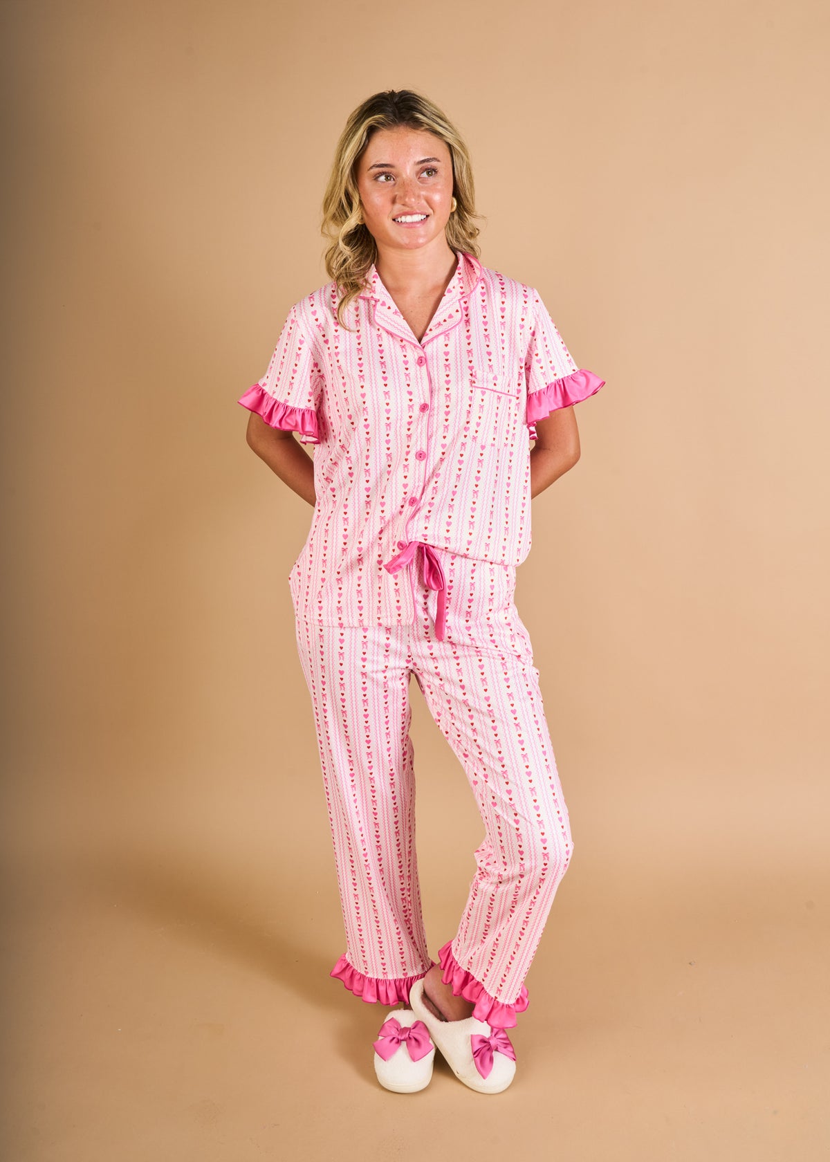 A person stands against a beige background wearing The Darling Effect’s Oh So Lovely Slumber Party Pajama Pants Set, crafted from ultra-soft fabric and showcasing pink stripes with delicate ruffle accents. They pair the set with white slippers adorned with bows. With blonde hair, they smile and look slightly to the side, hands in their pockets—making this gift-ready ensemble perfect for any special occasion.