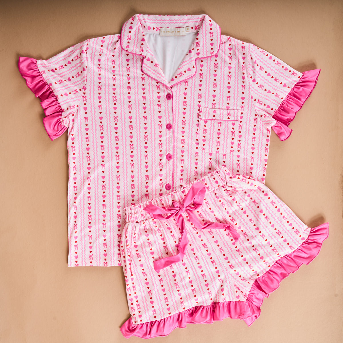 The Oh So Lovely Slumber Party Pajama Shorts Set by The Darling Effect features pink and white stripes, playful heart patterns, bright pink ruffle trim, and ultra-soft fabric. This short-sleeved set is laid flat on a beige background.